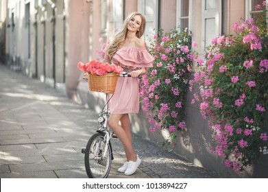 Pretty young woman is riding on a white bicycle in the city. She is smiling and wearing a pink off shoulder dress. On her bike is basket with pink roses.