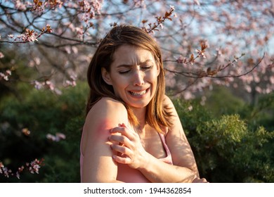 Pretty Young Woman Scratching Arm Beside Blooming Tree In Park In Spring. Redness And Itchy Skin As Allergic Reaction