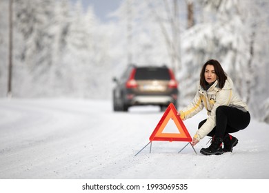 Pretty, young woman setting up a warning triangle and calling for assistance after her car broke down in the middle of nowhere on a freezing winter day