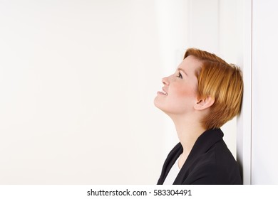 Pretty Young Woman With Short Red Hair And Rosy Cheeks Wearing Black Jacket, Leaning Back On White Wall Indoors And Looking Up. Side Portrait With Copy Space