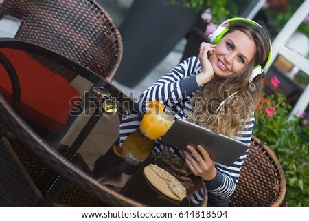 Pretty young woman sitting at the cafe and drinking lemonade. Beautiful woman with earphones and tablet listening the music
