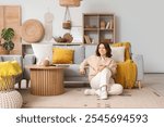 Pretty young woman sitting on floor near grey sofa in light living room