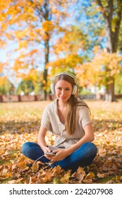 Pretty Young Woman Sitting On Grassy Ground With Dried Leaves  Listening Music On Ipod With Headset. Looking At Camera. Isolated On Nature Background.