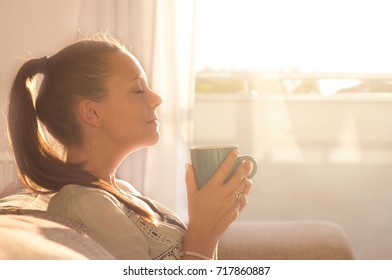 Pretty young woman sitting on sofa and enjoying first morning coffee on sunshine