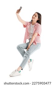 Pretty young woman with smartphone taking selfie while sitting on chair against white background