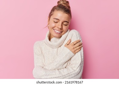 Pretty young woman smiles gently has closes eyes embraces herself feels comfort dressed in white knitted sweater isolated over pink background enjoys her beauty and youth. Love yourself concept