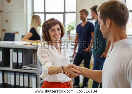 Pretty young woman smiling while greeting man with handshake, standing in office environment with other people in background soft focus