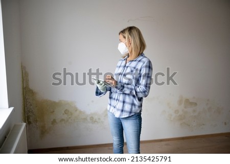 pretty young woman standing in front of wall with mold with tablet and mouth nose mask