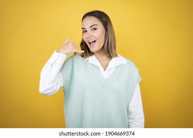 Pretty young woman standing over yellow background smiling doing phone gesture with hand and fingers like talking on the telephone