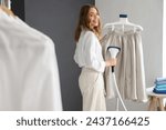 Pretty young woman steaming clothes with modern garment steamer in laundry room