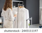 Pretty young woman steaming clothes with modern garment steamer in laundry room