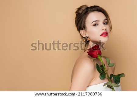 pretty young woman in strapless top holding red rose and looking at camera isolated on beige