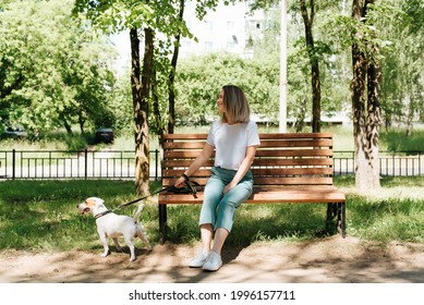 Pretty young woman in stylish clothes resting on a bench while walking the dog. Happy blonde girl and jack russell terrier on a walk in the park on a summer sunny day. - Shutterstock ID 1996157711