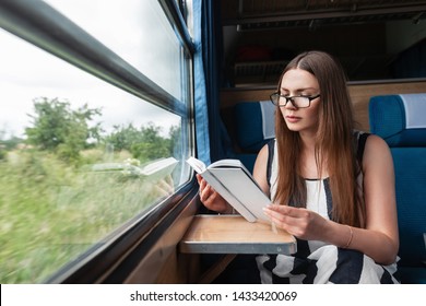 Pretty Young Woman In A Stylish Striped Summer Dress In Glasses Sits In The Train Near The Window And Reads An Interesting Book. Beautiful Girl Enjoys Reading. Euro-trip.