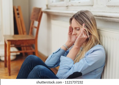 Pretty young woman suffering from a headache sitting against a warm radiator rubbing her temples with her fingertips - Shutterstock ID 1121737232