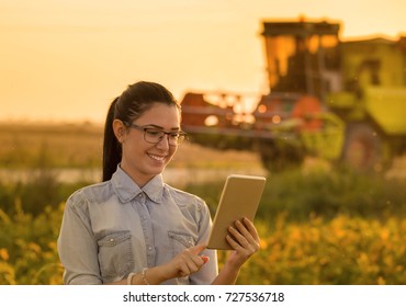 Pretty Young Woman With Tablet Standing In Soybean Field With Combine Harvester Working In Background