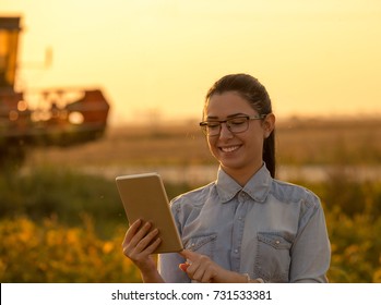 Pretty Young Woman With Tablet Standing In Soybean Field With Combine Harvester Working In Background