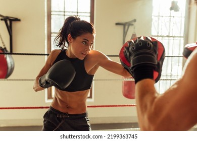 Pretty young woman trains in boxing ring with partner near red punching bag and other sparring equipment