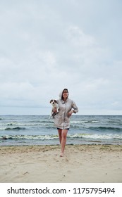 Pretty young woman in warm jacket holding adorable dog and looking at camera while standing on sand near beautiful waving sea on cloudy day