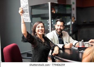 Pretty Young Woman Winning A Bingo Game And Raising Her Card While Playing With A Group Of Friends