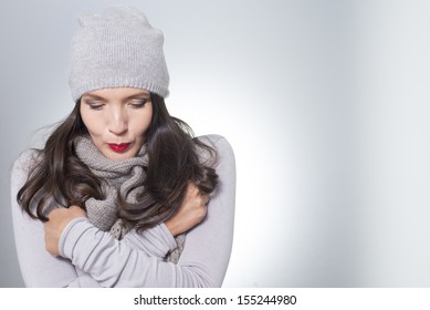 Pretty Young Woman In Winter Fashion Cuddling Down Inside Her Grey Woolly Knitted Jersey, Scarf And Cap On A Cold Day With Copyspace