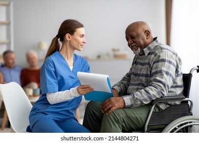 Pretty Young Woman In Workwear Nurse Helping Black Senior Man In Weheelchair Filling Papers, Cheerful Elderly African American Male Patient Signing Documents At Nursing Home