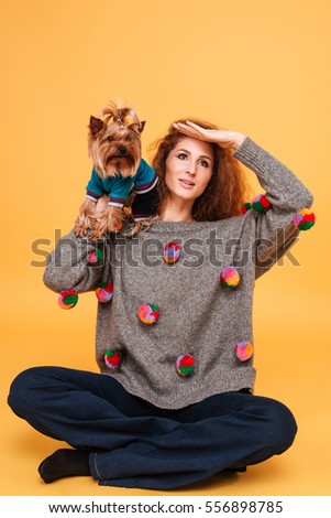 Pretty yuong woman with red hair sitting with her dog and looking far away isolated on orange background