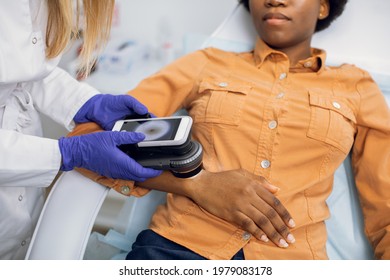 Preventive checkup, skin melanoma days concept. Close up of hands of female doctor dermatologist oncologist holding new generation dermatoscope, examining birthmarks and moles of afro lady