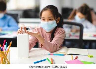 Preventive Measures Concept. Asian girl applying antibacterial hand sanitizer, pouring it on palms from the bottle, wearing disposable surgical face mask, sitting at desk in classroom during lesson - Shutterstock ID 1908796525