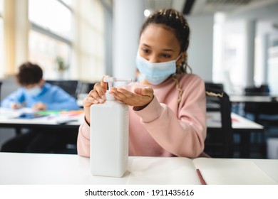 Preventive Measures Concept. Black girl applying antibacterial sanitizer, pouring it on palms from the bottle, wearing disposable surgical face mask, sitting at desk in classroom during lesson