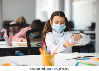 Preventive Measures. Small Girl Applying Antibacterial Sanitizer Spray On Hands, Wearing Disposable Protective Medical Mask, Sitting In Classroom At School. Stop The Spread Of Infection And Germs