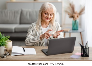 Preventive Measures, Stop Coronavirus Spread. Portrait of smiling mature woman spraying sanitizer on hand, selective focus. Happy senior lady using laptop, sitting at desk t home office - Powered by Shutterstock