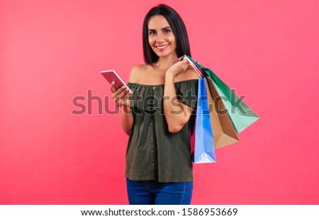 Prices slashed. Close-up photo of modern woman in a casual outfit, who is smiling at the camera, holding a smartphone in her right hand and three paper bags in her left hand.