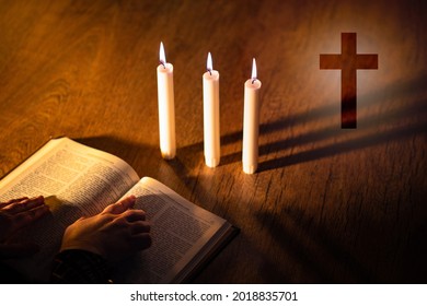 Priest hands on bible. Burning candles next to Christian cross. Cross symbolizes Catholicism or Orthodox. Attributes of Christianity on a wooden table. Catholic prays to God. Orthodox studying bible