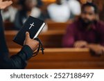 Priest is holding a bible and a rosary while preaching in a church with his congregation sitting in wooden pews in the background