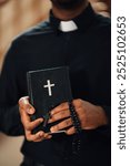 Priest holds a bible and rosary beads during a religious service in a church, exuding faith and devotion in a serene setting of worship and belief