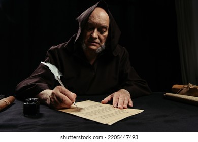 Priest In Hooded Robe Writing Chronicle On Parchment Isolated On Black