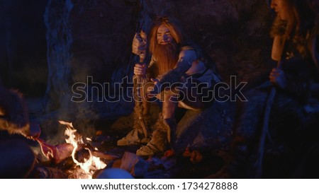Primal tribe of ancient wild people animal skin and stone axes eating frying meat on bonfire staying in cave. Neanderthals. Ice age.