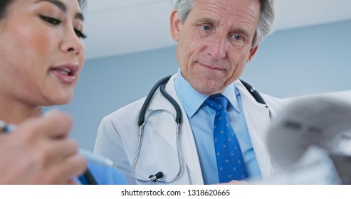 Primary Care Physician Talking To Surgeon About Patient Recovery. Medical Professionals Consulting Together On Paperwork. Senior Caucasian Male Doctor And Female Japanese Nurse