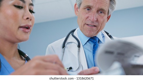 Primary Care Physician Talking To Surgeon About Patient Recovery. Medical Professionals Consulting Together On Paperwork. Senior Caucasian Male Doctor And Female Japanese Nurse