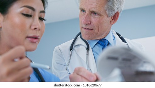 Primary Care Physician Talking To Surgeon About Patient Recovery. Medical Professionals Consulting Together On Paperwork. Senior Caucasian Male Doctor And Female Japanese Nurse