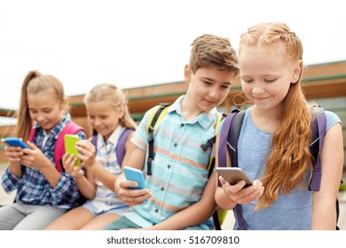 primary education, friendship, childhood, technology and people concept - group of happy elementary school students with smartphones and backpacks sitting on bench outdoors - Powered by Shutterstock