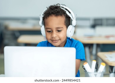 Primary school boy student with headphones using laptop, typing on keyboard, sitting in classroom interior - Powered by Shutterstock