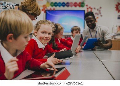 Primary School Children Are In The Classroom, Learning About And Using Digital Tablets. One Of The Girls Is Smiling At The Camera.