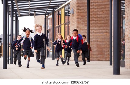 Primary School Kids, Wearing School Uniforms And Backpacks, Running On A Walkway Outside Their School Building, Front View