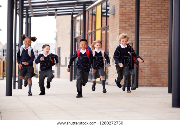 Primary\
school kids, wearing school uniforms and backpacks, running on a\
walkway outside their school building, front\
view