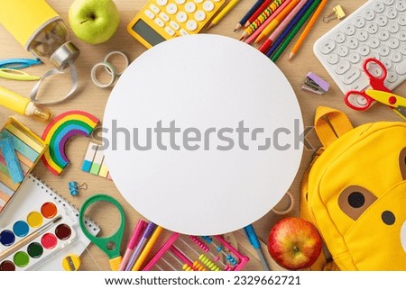 Primary school setup. Top view of assorted stationery, album, paint palette, pencils, keyboard, calculator, bear backpack, water bottle, apples and more on wooden desk with empty circle for advert