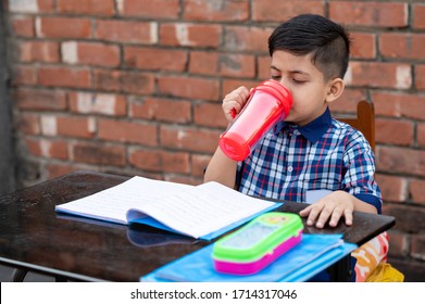 Primary School Student Drinking Water With Red Water Bottle While Sitting In Class On Study Table. Indian School Student In Classroom.