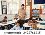 Primary school teacher walking in classroom while multiethnic students studying. Male teacher holding textbook helping schoolchildren during class. Professional educator walking in classroom.