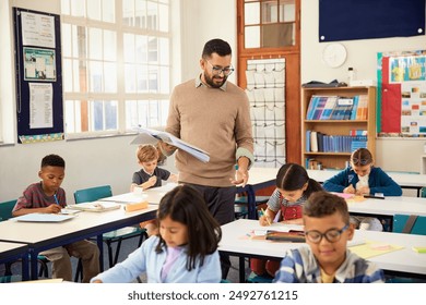 Primary school teacher walking in classroom while multiethnic students studying. Male teacher holding textbook helping schoolchildren during class. Professional educator walking in classroom. - Powered by Shutterstock